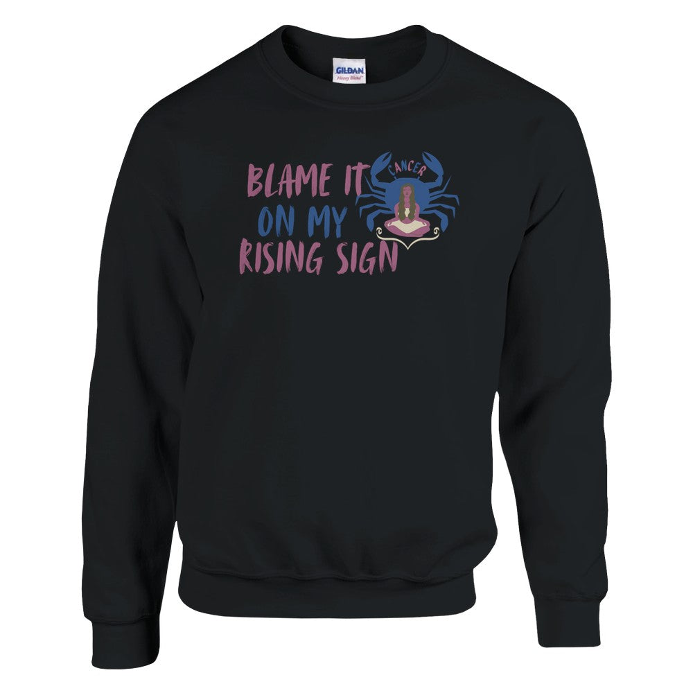 Black sweatshirt with a Cancer zodiac-themed design featuring the phrase 'Blame It On My Rising Sign.' The design includes a colorful crab illustration with the word 'Cancer' above it and an abstract depiction of a person in a relaxed pose, conveying a playful astrology-inspired theme.