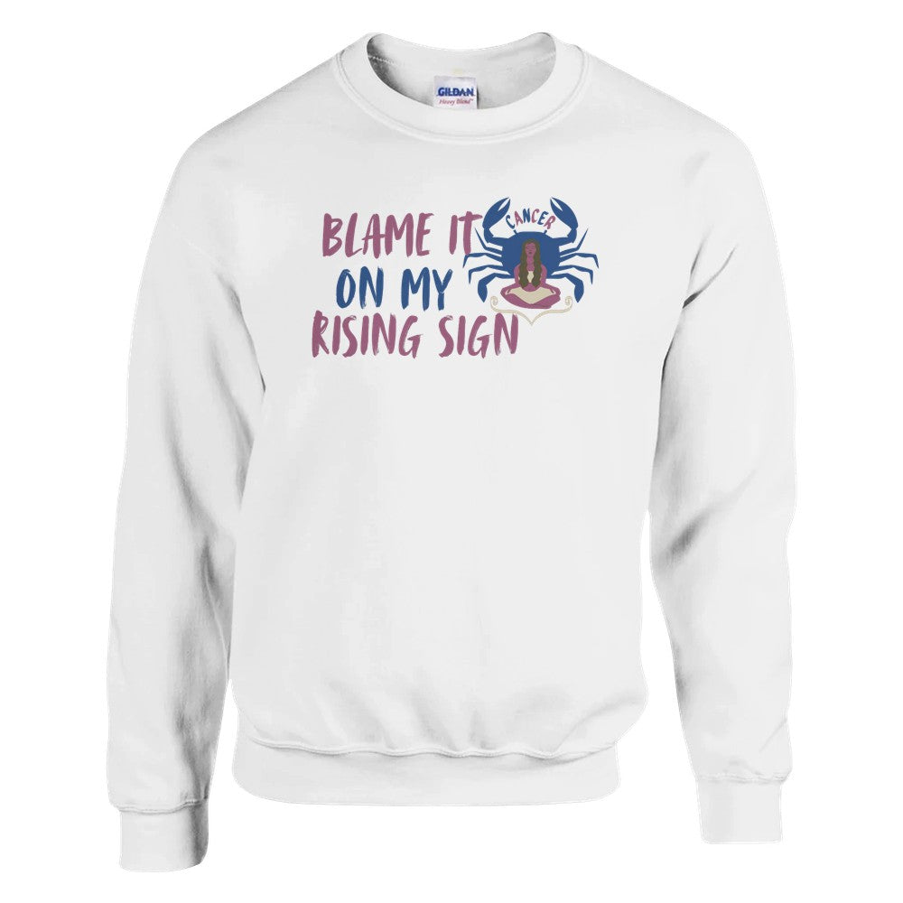 White sweatshirt with a Cancer zodiac-themed design featuring the phrase 'Blame It On My Rising Sign.' The design includes a colorful crab illustration with the word 'Cancer' above it and an abstract depiction of a person in a relaxed pose, conveying a playful astrology-inspired theme.