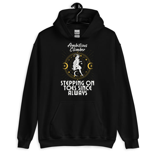 Front of black Capricorn-themed hoodie with retro design, featuring text 'Ambitious Climber' above an illustration of a determined figure, surrounded by celestial symbols, and 'Stepping on Toes Since Always' below.