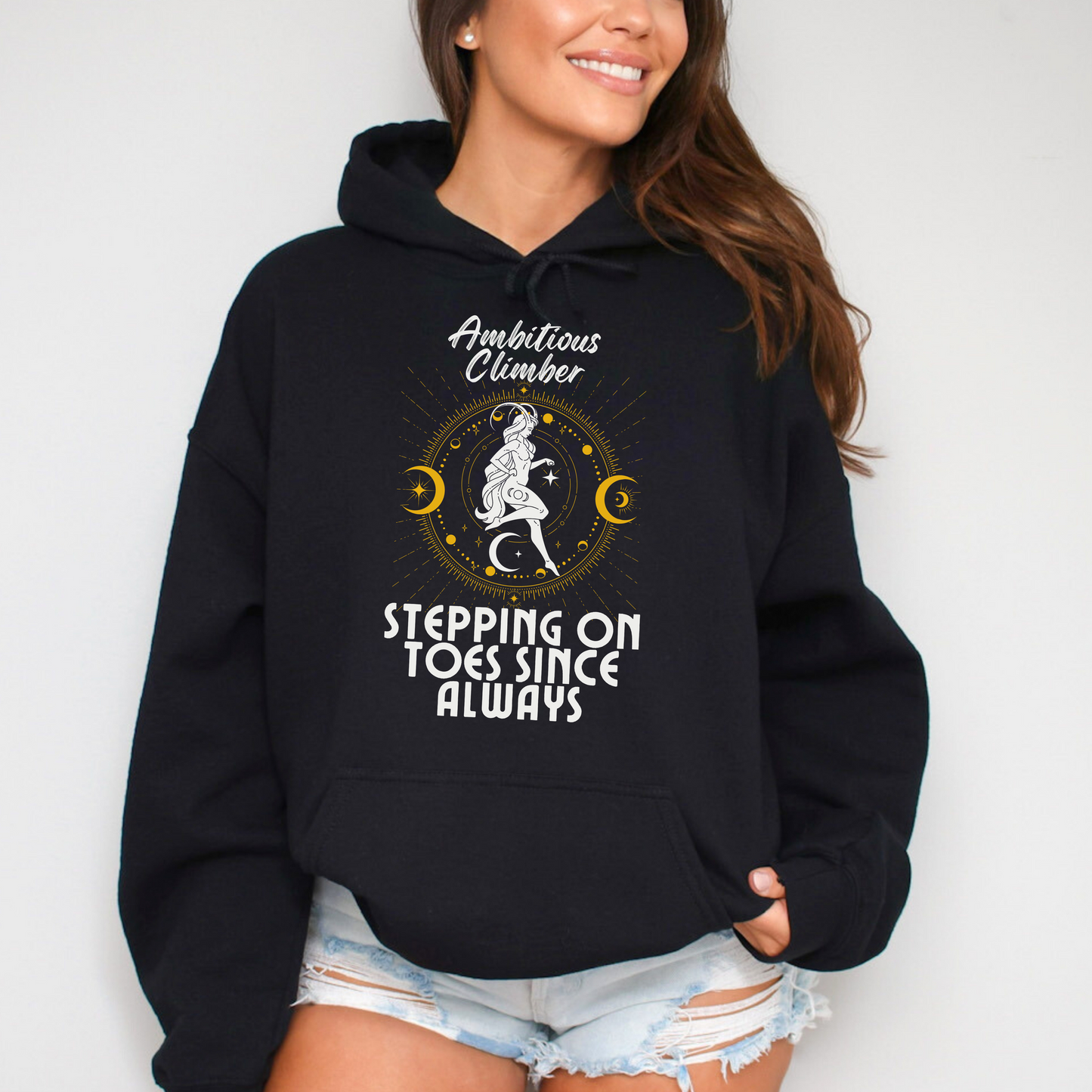 Front of black Capricorn-themed hoodie with retro design, featuring text 'Ambitious Climber' above an illustration of a determined figure, surrounded by celestial symbols, and 'Stepping on Toes Since Always' below.