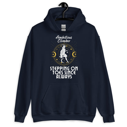 Front of navy blue Capricorn-themed hoodie with retro design, featuring text 'Ambitious Climber' above an illustration of a determined figure, surrounded by celestial symbols, and 'Stepping on Toes Since Always' below.