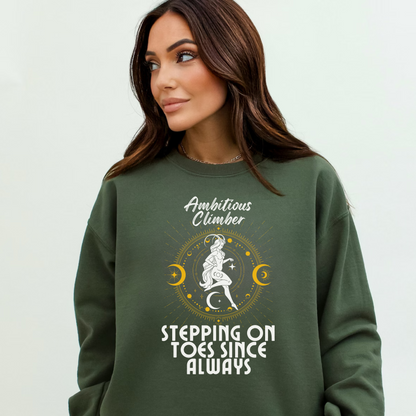 Front of military green Capricorn-themed sweatshirt with retro design, featuring text 'Ambitious Climber' above an illustration of a determined figure, surrounded by celestial symbols, and 'Stepping on Toes Since Always' below.