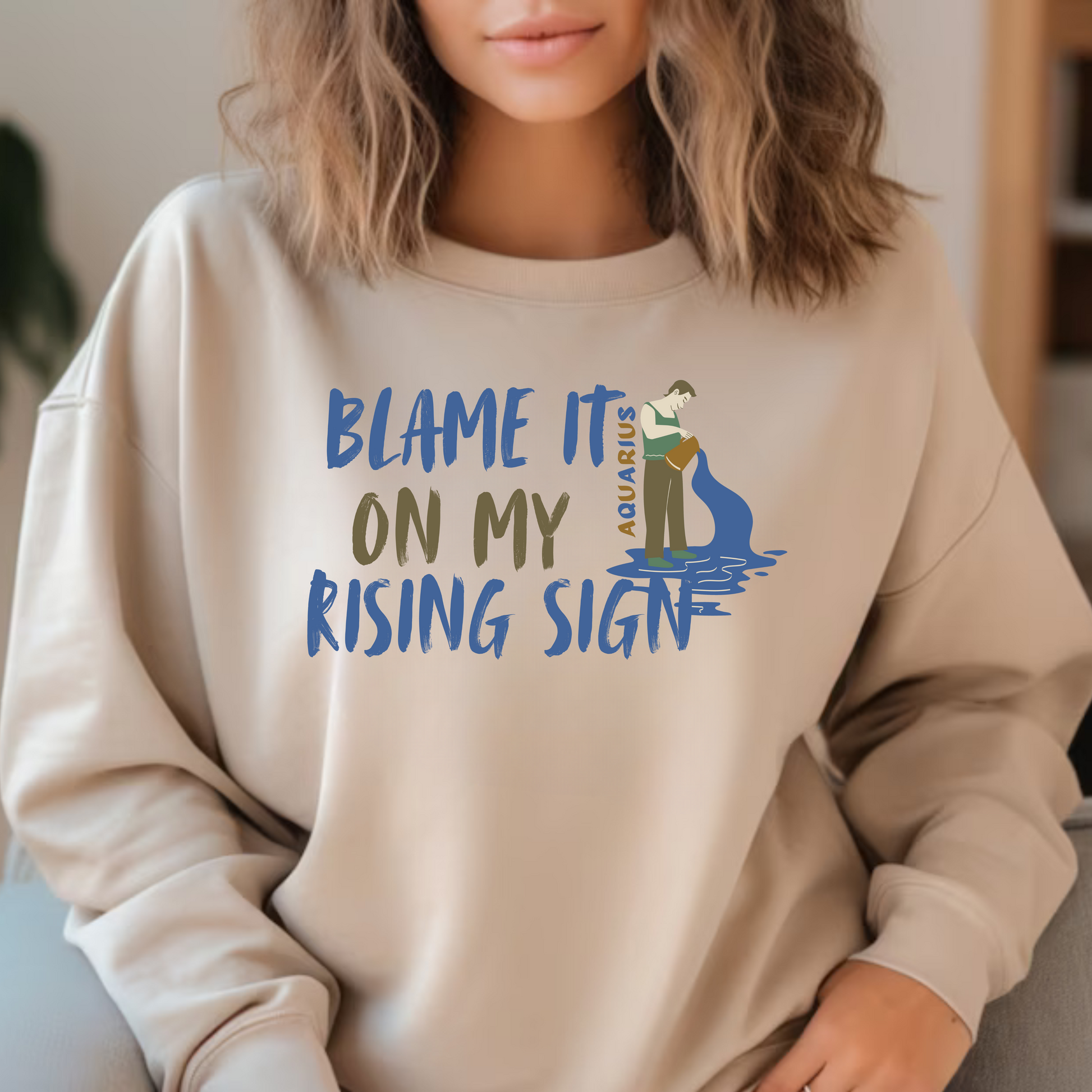 Sand Aquarius sweatshirt with 'Blame It On My Rising Sign' text and colorful Aquarius water-bearer illustration in blue and brown tones.