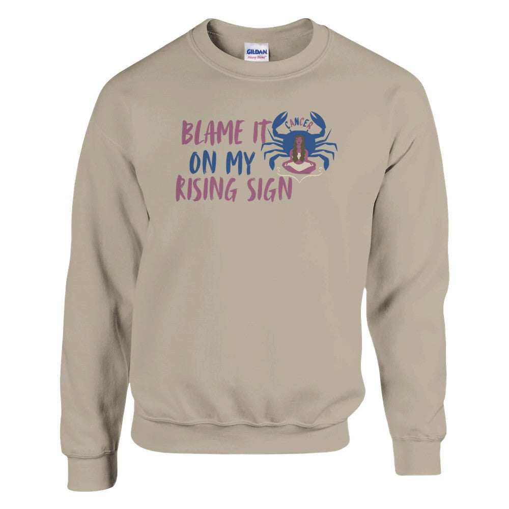 Sand sweatshirt with a Cancer zodiac-themed design featuring the phrase 'Blame It On My Rising Sign.' The design includes a colorful crab illustration with the word 'Cancer' above it and an abstract depiction of a person in a relaxed pose, conveying a playful astrology-inspired theme.